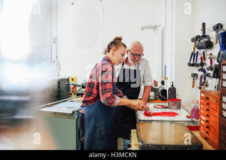 Artesano Senior/técnico supervisar joven como ella usa rodillo de tinta para la impresión en serigrafía taller de artes del libro