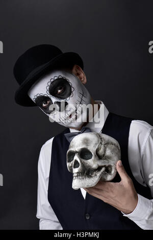  Retrato de un hombre joven con calaveras mexicanas maquillaje, vestidos de pajarita y sombrero, sosteniendo un cráneo en su mano junto a su Fotografía de stock