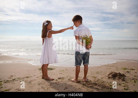 Niño sosteniendo flores detrás de su espalda, de pie junto a la niña en la playa