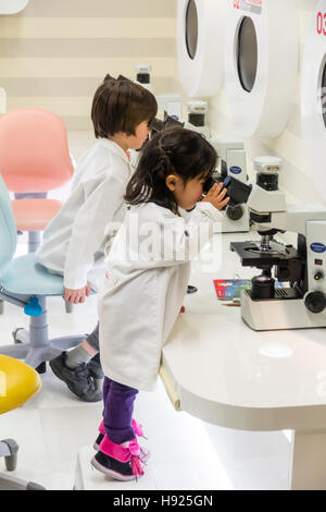 Japón, Nishinomiya, KidZania. Los niños, niño y niña en batas de laboratorio blancas usando microscopios, niña de puntillas fatigar para ver. Foto de stock