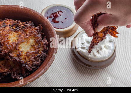 Latkes en un cuenco de cerámica marrón rústico con crema agria y aparte latkes mermelada de fresa con crema agria en inmersión con la mano Foto de stock
