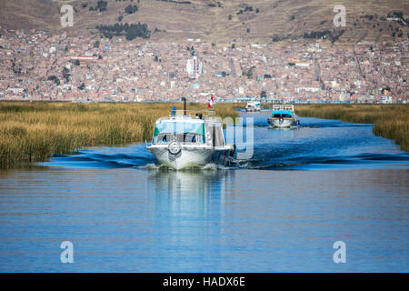 Los barcos turísticos en el Lago Titicaca, flanqueado por campos de juncos de totora, la ciudad de Puno, en el fondo, Puno, Perú Foto de stock