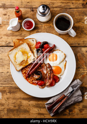 Completo desayuno inglés con huevos fritos, salchichas, tocino, frijoles, tostadas, tomates y champiñones sobre fondo de madera Foto de stock