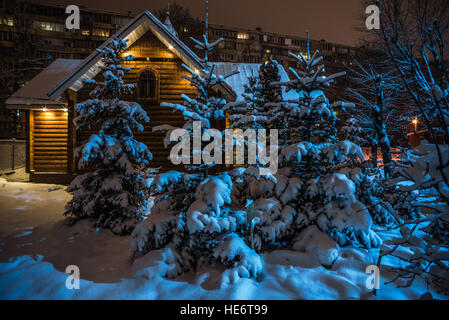 Pequeña cabaña y arbustos cubiertos de nieve, sobre un fondo de edificio de gran altura en la noche Foto de stock