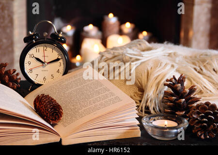 La lectura de un libro de bolsillo en la noche a la luz de las velas junto a una chimenea abierta en una acogedora casa inglesa con una manta (lanzar) en invierno Foto de stock