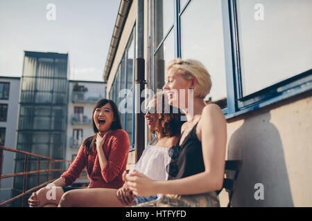 Disparo de smiling amigas jóvenes sentados juntos en la terraza. Las mujeres multirraciales relajarse al aire libre y divertirse.