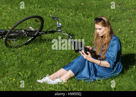 Mujer y vintage bicicleta, portátil, césped verde, verano. Cabello rojo chica sentada en el césped de la escuela, la una tableta, vieja bicicleta retro ba Fotografía de stock -