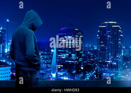 Hacker silueta de hombre encapuchado parado en la cima del edificio por la noche Foto de stock