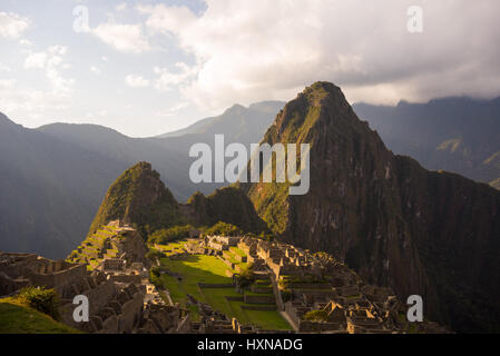 Machu Picchu parcialmente iluminada por los últimos rayos del sol. Amplio ángulo de visión de las terrazas con vistas panorámicas del cielo y resplandeciente de sendero en primer plano. Foto de stock