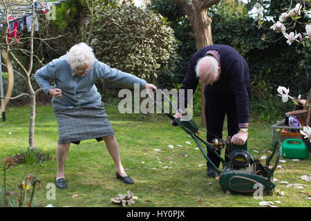 Los pensionistas ancianos intentando iniciar un cortacésped de gasolina en un inglés jardín residencial en una tarde de primavera, Surrey, Inglaterra, Reino Unido Foto de stock
