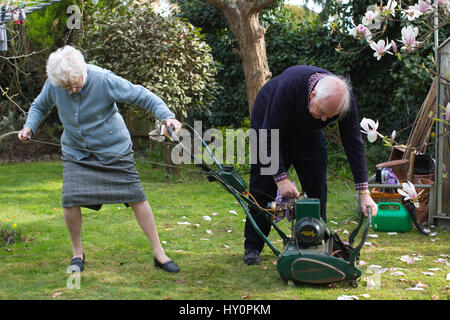 Los pensionistas ancianos intentando iniciar un cortacésped de gasolina en un inglés jardín residencial en una tarde de primavera, Surrey, Inglaterra, Reino Unido Foto de stock