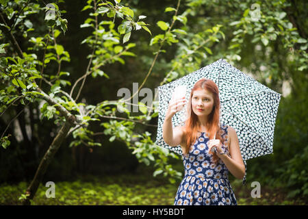 Finlandia, Pirkanmaa, Tampere, mujer vistiendo vestido floral de pie con paraguas en el parque y teniendo selfie