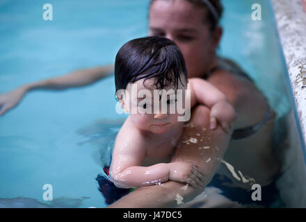 Triste niñito gordito con mejillas aferrado al brazo de su madre en una piscina azul vistiendo un traje de baño.