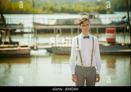 Joven hombre flaco, elegantemente elegantemente vestido con camisa blanca, pantalón  gris con tirantes y pajarita. Retrato joven en el muelle en el fondo del r  Fotografía de stock - Alamy
