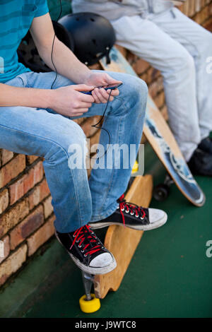 Dos muchachos adolescentes sentados en un muro de ladrillo, con skateboards, escuchando música Foto de stock