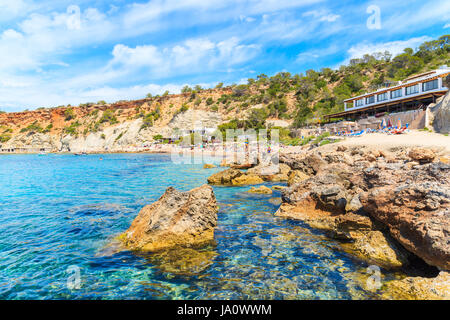 Vista de la playa de Cala d'Hort con piedras en el agua del mar y el restaurante en el fondo, la isla de Ibiza, España Foto de stock