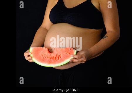Una mujer embarazada sosteniendo una rebanada de sandía con una mordida fuera de ella, lo que demuestra su ombligo. Foto de stock