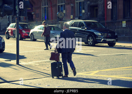 Hombre de negocios turista con maletas trolley maletín con ruedas en las calles de Glasgow Crossing Road hasta el destino o el hotel Foto de stock