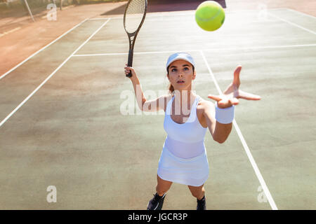 Hermosa joven jugador de tenis femenino sirviendo la bola. Mujer joven en ropa deportiva jugando tenis en la cancha. Foto de stock