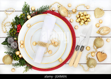 Cena de Navidad cubierto con placas de porcelana, decoraciones de adornos de oro, acebo, muérdago, hiedra y abeto blanco angustiado sobre fondo de madera Foto de stock