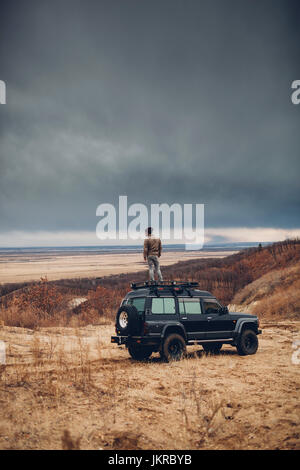 Hombre distante de pie en la parte superior del vehículo utilitario deportivo sobre la tierra contra el cielo nublado, Amur, Rusia