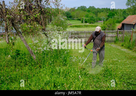 Hombre utilizando un local strimmer para cortar el césped largo en el jardín de una casa rural en una aldea en el condado de Zala hungría