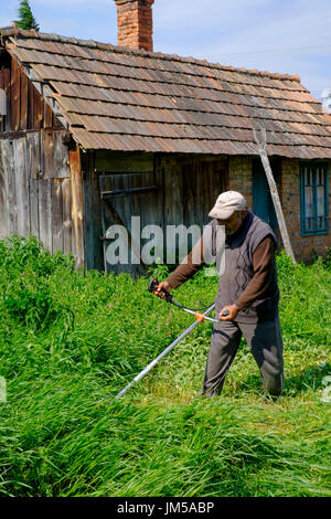 Hombre utilizando un local strimmer para cortar el césped largo en el jardín de una casa rural en una aldea en el condado de Zala hungría