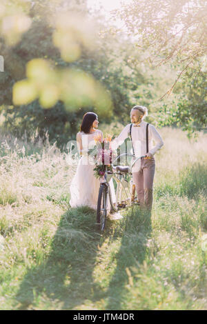 La moderna vestida de recién casados están llevando la bicicleta con flores rosas y paseos en el verde bosque soleado.