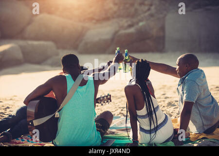 Los jóvenes brindando con botellas de cerveza mientras está sentado en la playa. Grupo de amigos tener bebidas juntos.