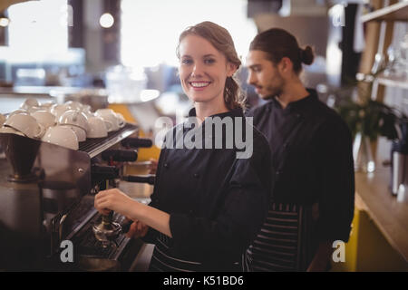 Retrato de mujer sonriente barista con cafetera expreso contra el camarero en la cafetería Foto de stock