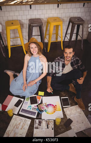 Ángulo alto retrato de profesionales creativos sonriente joven sentada en el piso con hojas en la cafetería Foto de stock