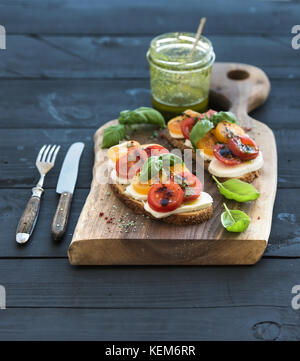 Mozzarella, tomate y albahaca sándwiches en una tabla de cortar de madera oscura, pesto jar, vajillas sobre fondo negro