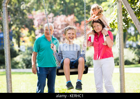 Feliz nieto kid divirtiéndose en swing con viejos abuelos de pie detrás en el parque