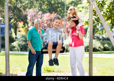 Niño feliz divirtiéndose en swing con abuelos de pie detrás en el parque