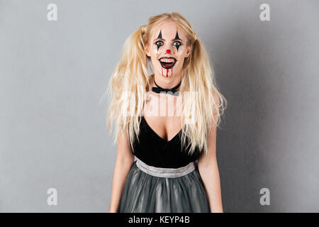 Retrato de una mujer rubia Risa loca en halloween maquillaje de payaso y vetas de sangre mirando a la cámara aislada sobre fondo gris Fotografía de stock