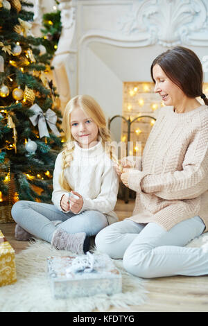Senior femenino en el sweater y jeans de trenzado del cabello de su nieta por arbol de navidad Foto de stock