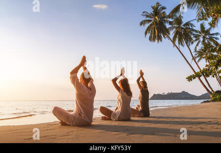 Grupo de tres personas que practican yoga postura del loto en la playa para el relax y el bienestar, cálido paisaje de verano tropical con palmeras