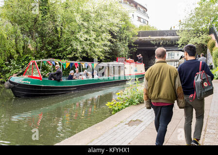 Narrowboat o barcaza pasando sobre el Regents Canal con gente caminando en el camino de sirga canalside cerca de la ciudad de Camden, Londres, Inglaterra, Reino Unido.