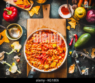 Chili con carne plato vegetariano en pan sobre la tabla de cortar de madera con especias y verduras ingredientes para cocinar en la cocina oscura de fondo de la tabla, de arriba vi Foto de stock
