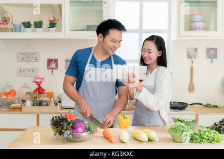 Joven y bella asiática el esposo y la esposa buscando receta de menú en digital tablet en la cocina en casa. feliz pareja el amor y el concepto de tecnología. Foto de stock
