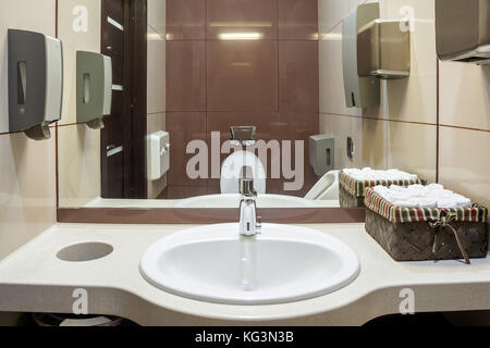 Lavabo blanco grande en baño público con toallas de papel, el secador de  manos automático, lavabo, fregadero seca marrón con paredes de azulejos, en  el fondo un wc Fotografía de stock 