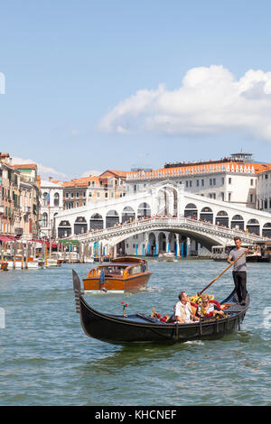 Una pareja de turistas de remo de gondolero en su góndola delante del puente de Rialto, Venecia, Italia con un taxi acuático pasando por Foto de stock
