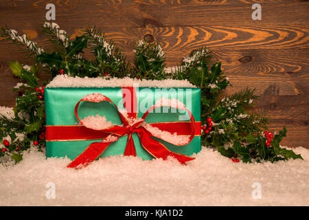 Regalo de navidad regalo envuelto en papel verde con una cinta roja con el acebo y la nieve contra un fondo de madera, espacio para copiar su propio mensaje. Foto de stock