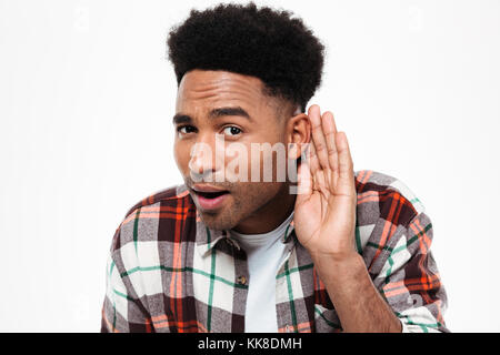 Close Up retrato de un curioso afro americana hombre intentando escuchar algo mientras mantiene la mano en la oreja aislado sobre fondo blanco. Foto de stock