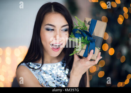 Emocionados mujer en la noche de Navidad sentado cerca de árbol y levantando su regalo de Navidad para escuchar lo que hay en su interior