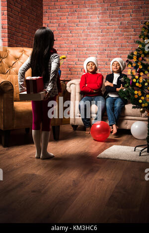 Cute little indian Chica sujetando u ocultar la sorpresa de regalo de navidad con ambas manos detrás de su cuerpo y el pequeño hermano sentado en el sofá con los ojos cerrados