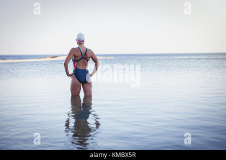 Nadadora de agua abierta femenina determinada y pensiva con las manos en las caderas en las olas del océano Foto de stock