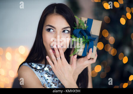 Emocionados mujer en la noche de Navidad sentado cerca de árbol y levantando su regalo de Navidad para escuchar lo que hay en su interior
