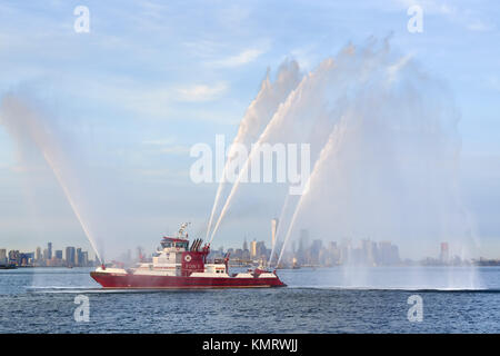 Fire Fighter II fireboat con salpicaduras de agua a plena potencia en la parte delantera del bajo Manhattan (incluyendo uno del World Trade Center) al atardecer. El puerto de la ciudad de Nueva York Foto de stock