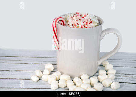 Bebida de chocolate caliente en taza blanca con malvaviscos y crema batida con bastón de caramelo de Navidad en madera blanqueada Foto de stock